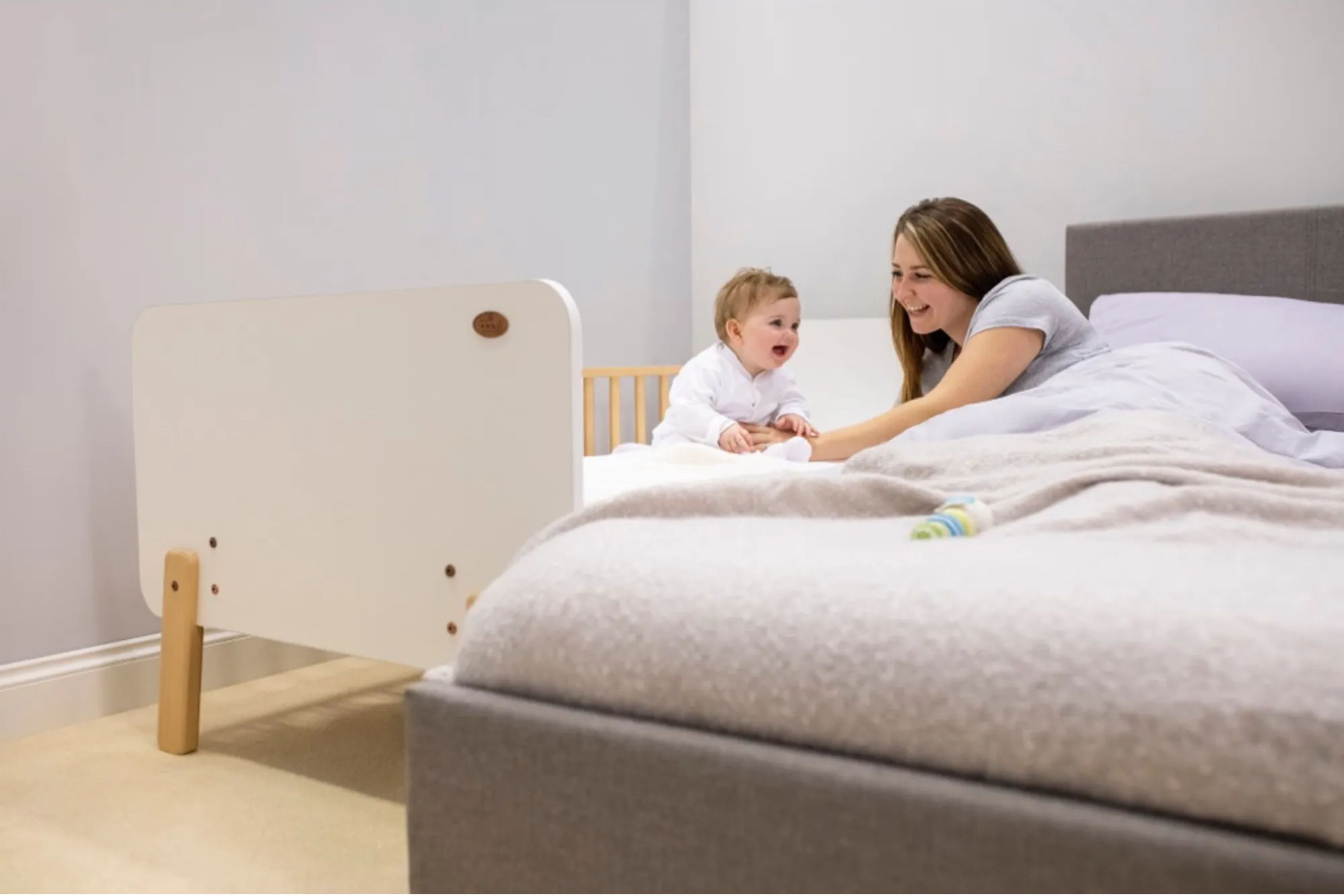 A smiling mother sits beside her baby who is comfortably lying in the Boori Natty Bedside Bed, placed next to her bed for easy access and closeness