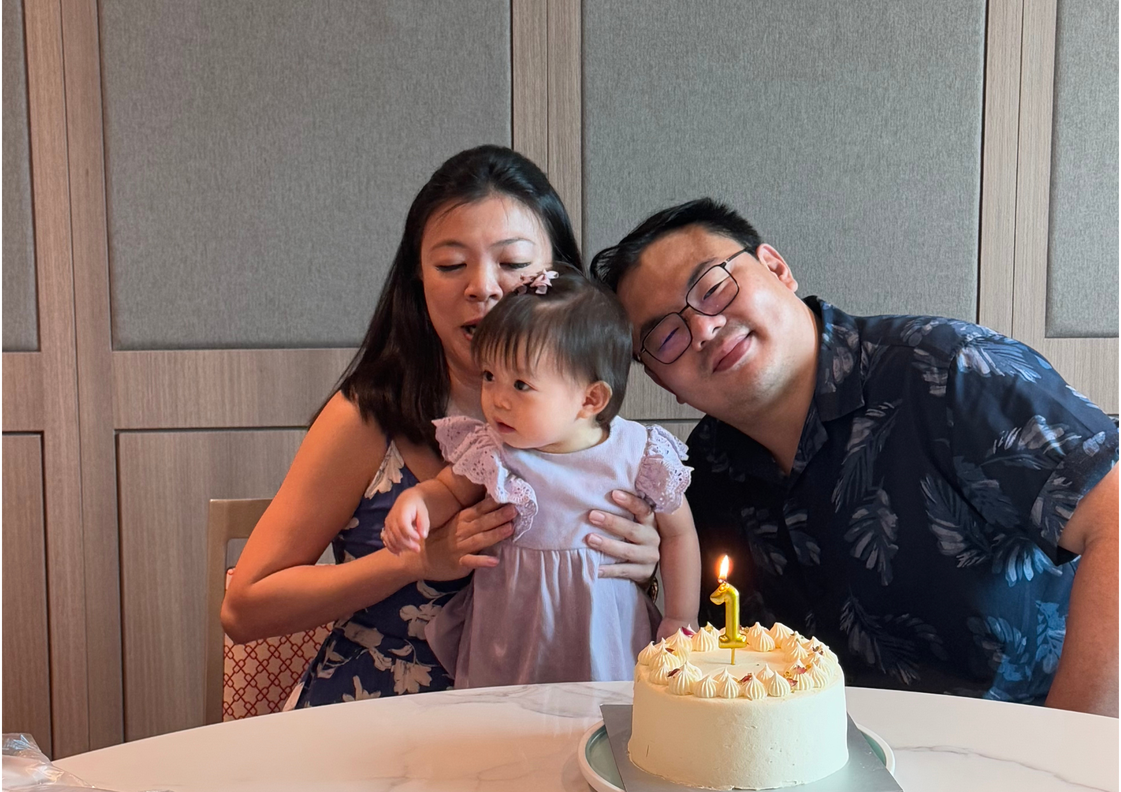 Family of three with a baby and a birthday cake with a lit candle indoors.