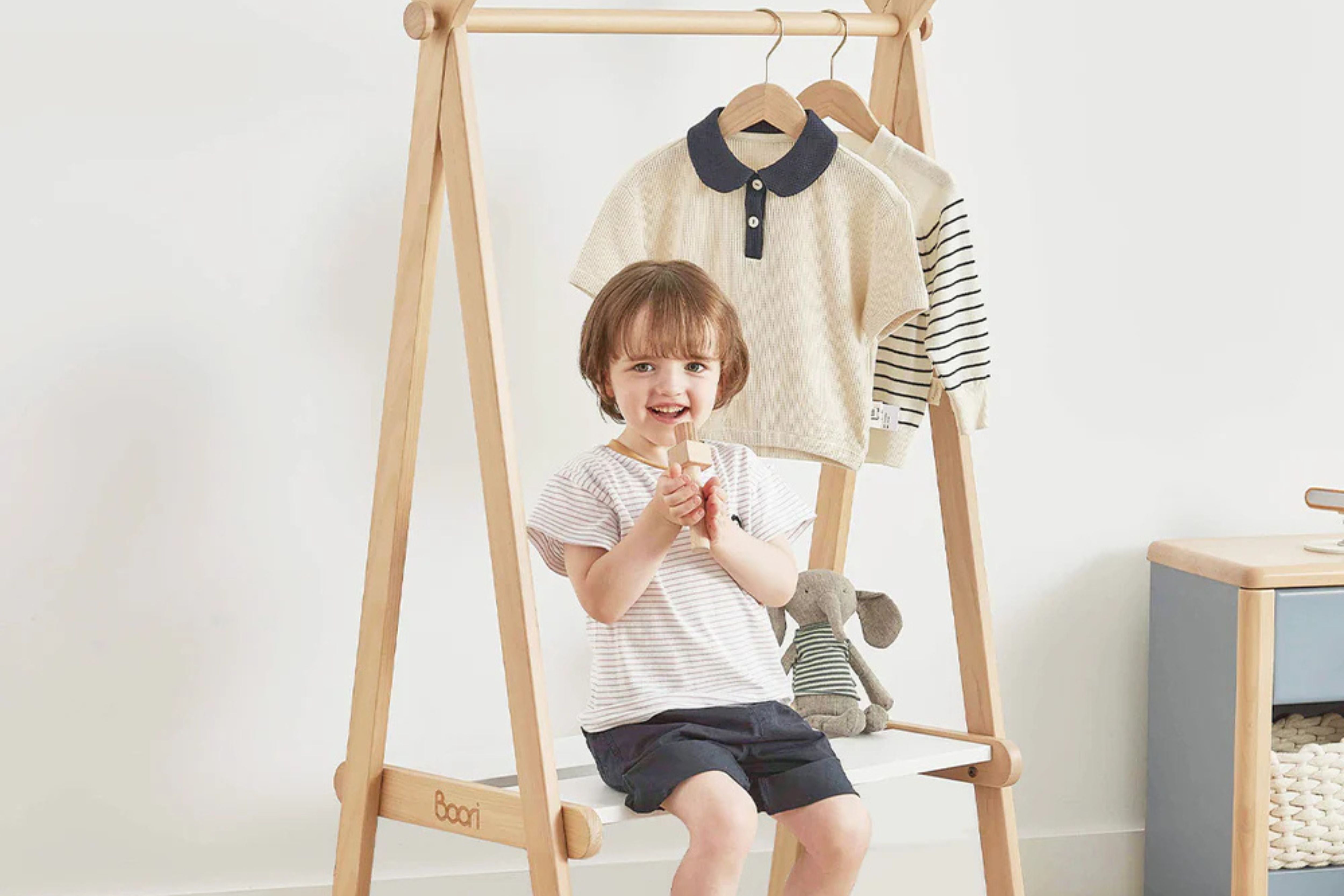Child sitting on the lower shelf of the Boori Forest Clothing Rack, with clothes hanging from the top rail above