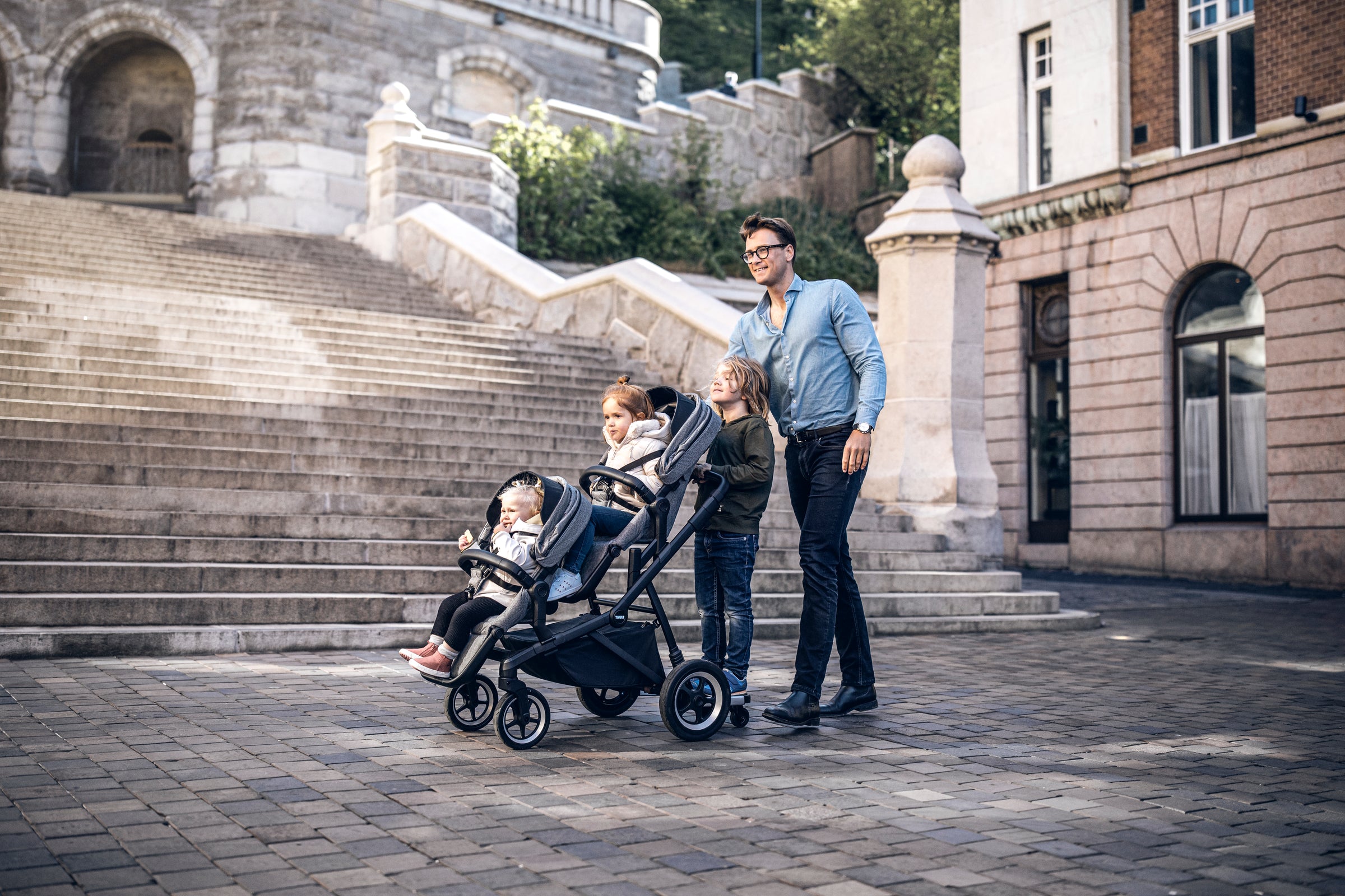 Dad pushing children in double stroller with rider board attached