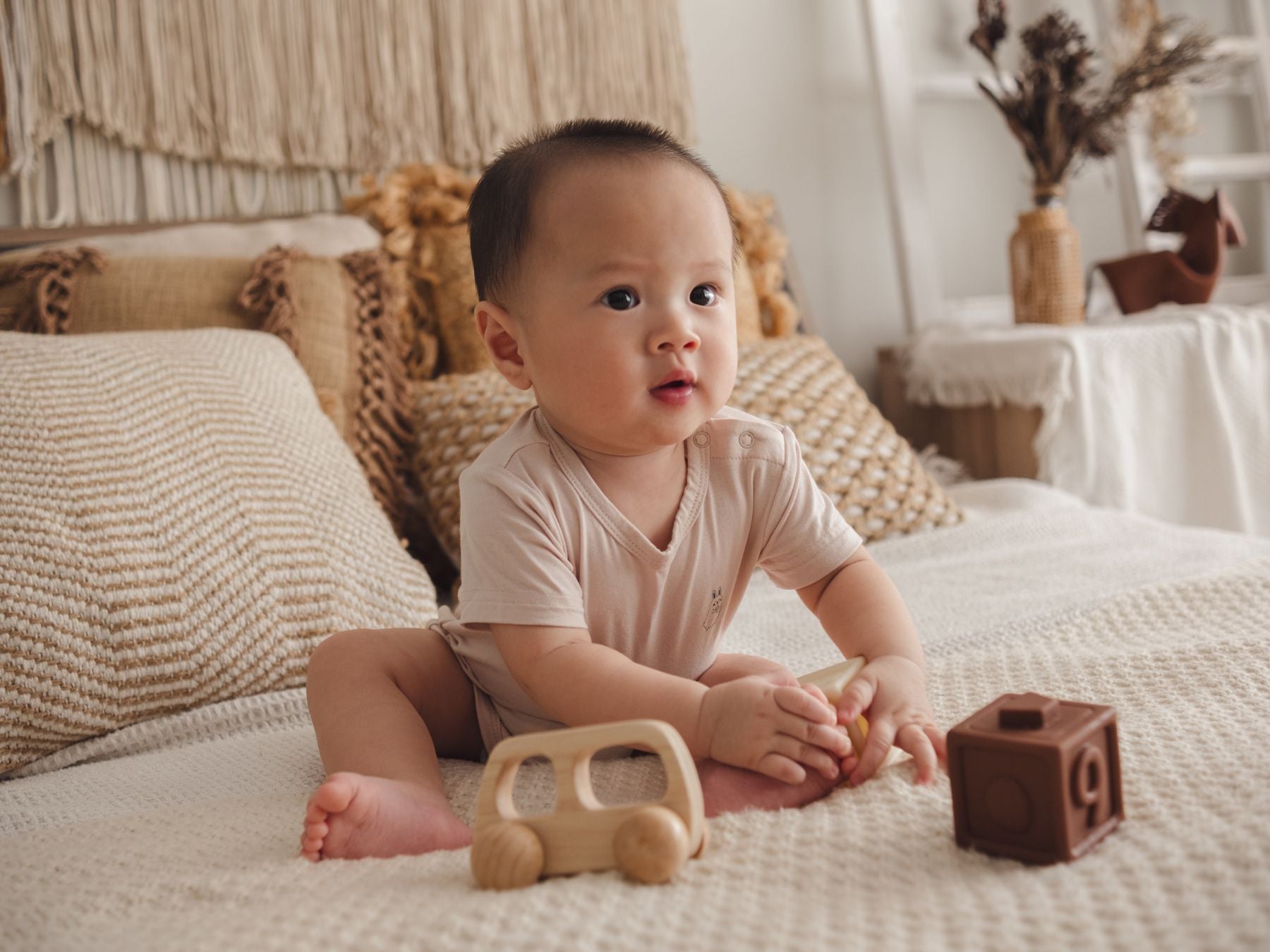 Baby in a beige onesie sitting on a bed, playing with toys in a cosy bedroom