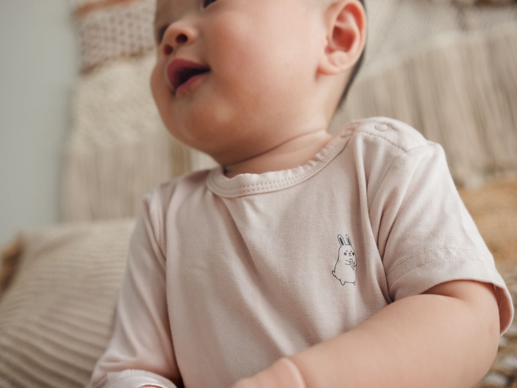 Baby sitting upright on a bed in a beige onesie