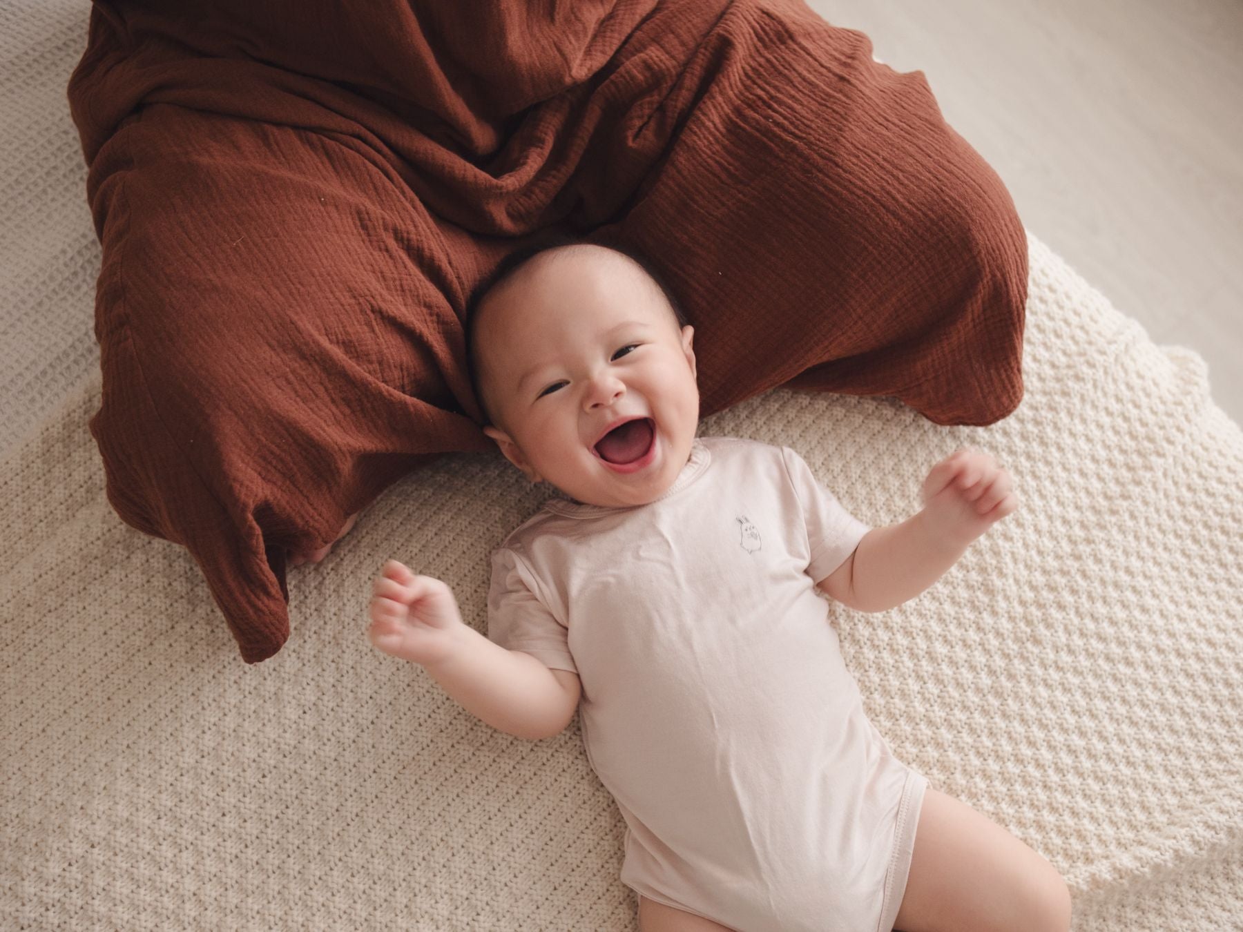 Happy baby lying on mum’s lap, wearing a beige onesie and smiling brightly