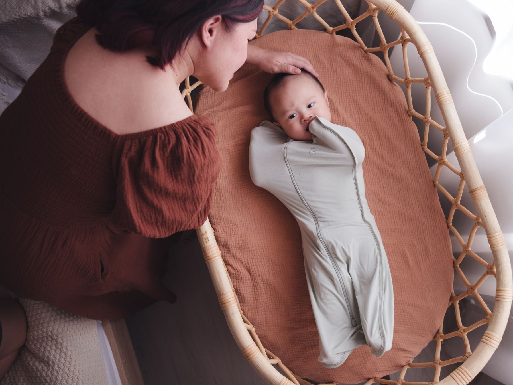 Woman leaning in to pat her baby’s head as the baby rests in a cot in a sage swaddle bag