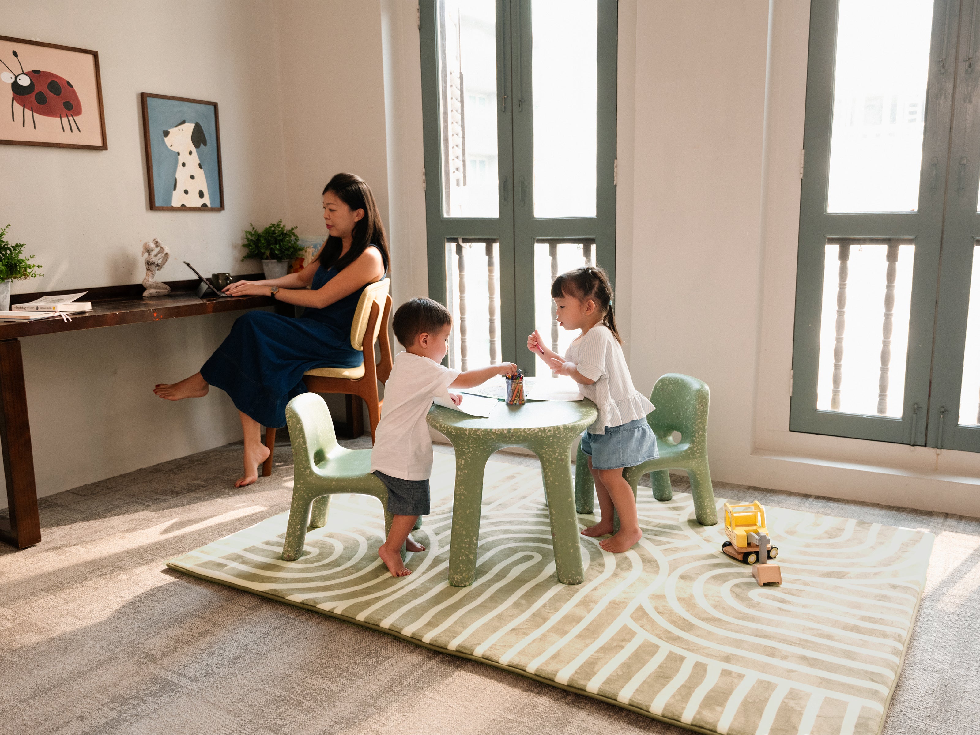 A woman sitting in a study corner while two toddlers play nearby on the Lollibly Kaya Plush Playmat.