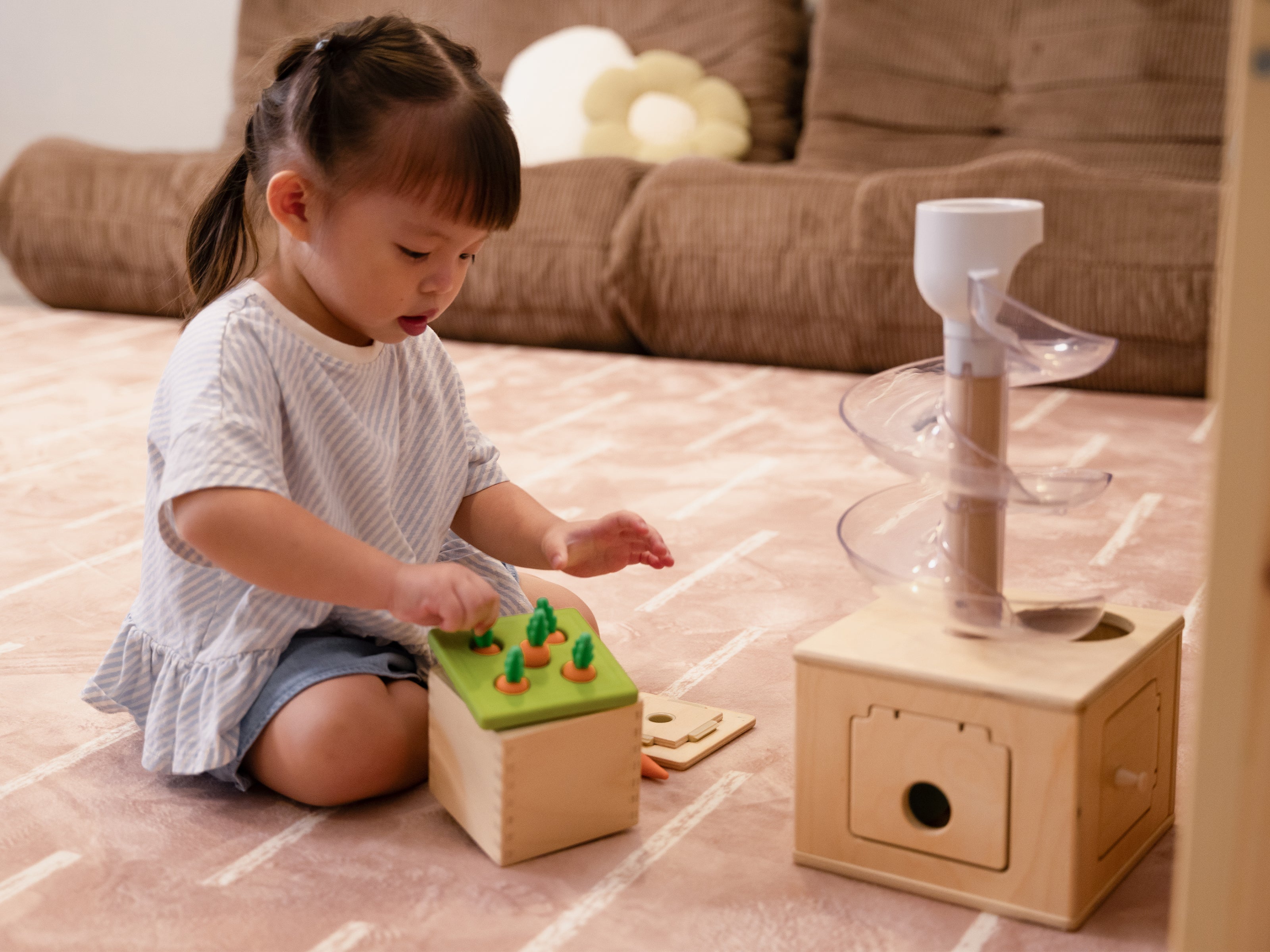 A young child sitting on the Lollibly Shortbread Plush Playmat, featuring a patterned design, while playing with wooden toys in a cosy living room.
