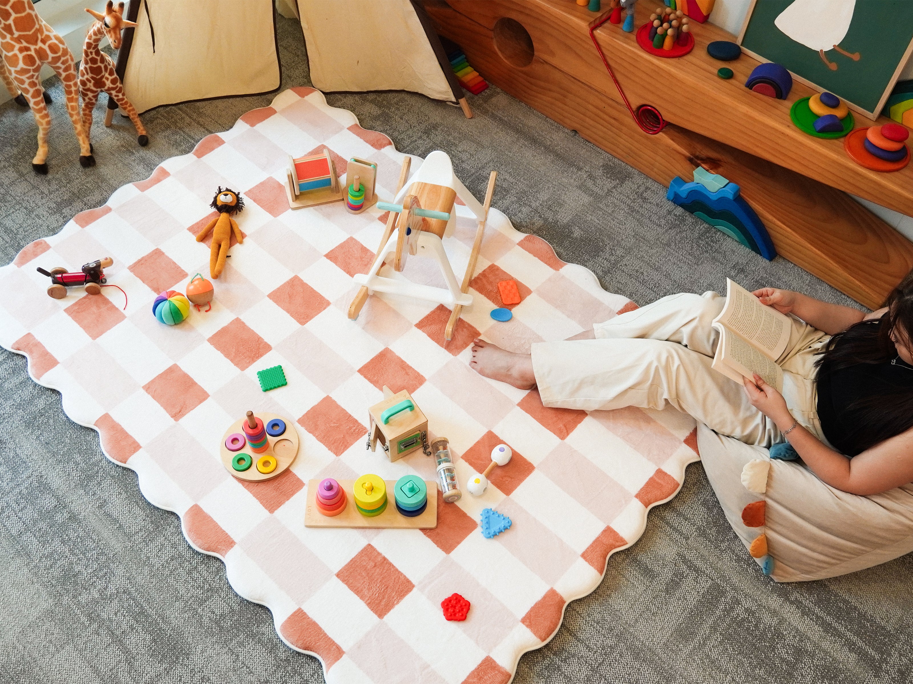 A person sitting among scattered toys on the Lollibly Waffle Plush Playmat, featuring a checkered pattern and scalloped edges in a cosy playroom.